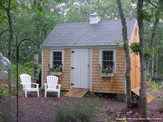 a small wooden shed sitting in the middle of a forest with two white chairs next to it