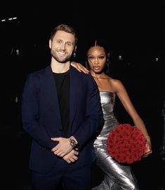 a man and woman standing next to each other in front of a black background holding roses