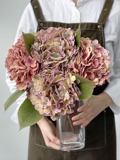 a woman in an apron holding a vase with pink flowers inside it and green leaves on the side