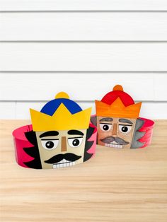 two paper crowns sitting on top of a wooden table next to each other with faces painted on them