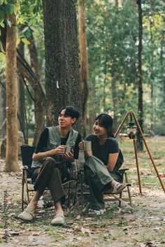 two people sitting in chairs next to each other near a tree and an easel