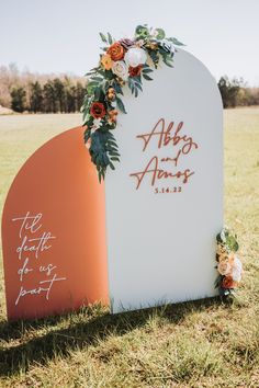 an orange and white sign with flowers on it in the middle of a grassy field