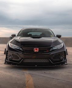 the front end of a black car parked on top of a parking lot with cloudy skies in the background