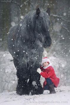 a small child is standing next to a large horse in the snow with it's head down