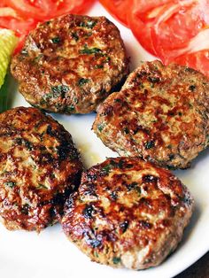 four hamburger patties on a white plate with corn and tomatoes in the background,