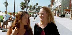 two women standing next to each other in front of palm trees and people on the sidewalk