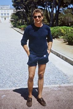 a man standing in front of some trees wearing shorts and sandals with his hands on his hips