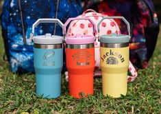 three insulated travel mugs sitting in the grass next to a back pack and duffel bag