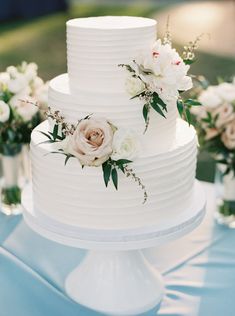 a three tiered white cake with flowers on the top is sitting on a blue tablecloth