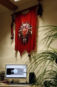 a desk with a computer, monitor and plant in front of a red cloth hanging on the wall