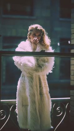 a woman in a fur coat standing on a balcony looking at the camera and talking on her cell phone