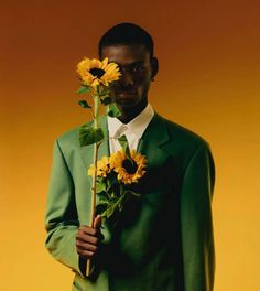 a man in a green suit holding sunflowers on his shoulder and looking at the camera