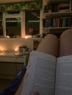 someone is reading a book in their bed with candles on the window sill behind them