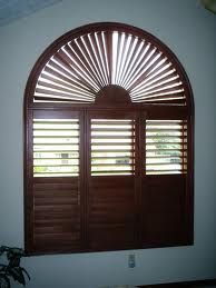 an arched window with wooden shutters in a white walled room, and a potted plant next to it