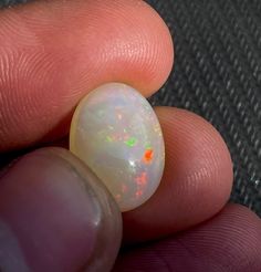 a hand holding a white opal in it's left thumb, with an orange and green speckled center