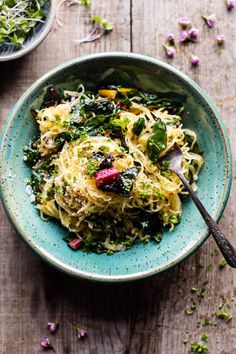 a blue bowl filled with pasta and vegetables on top of a wooden table next to a spoon