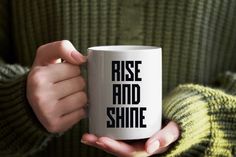 a person holding a white coffee mug with the words rise and shine on it in black