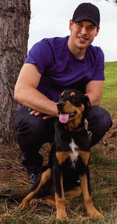 a man kneeling down next to a dog in the grass near a tree with his tongue hanging out