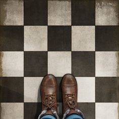 a pair of brown shoes sitting on top of a checkerboard floor next to a person's legs