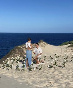 a man and woman sitting on the sand next to some white flowers in front of an ocean