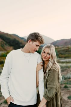 a man and woman standing next to each other in front of the mountains at sunset