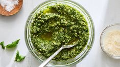 a glass bowl filled with pesto next to two wooden spoons on top of a white table