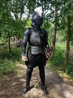 a man dressed in armor standing on a dirt path next to some trees and bushes