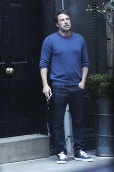 a man standing in front of a black door with his hands in his pockets and looking at the camera