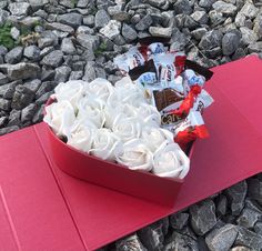 a heart shaped box filled with white roses on top of a red mat next to rocks