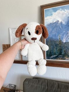 a hand holding a stuffed dog in front of a painting on the wall with mountains behind it