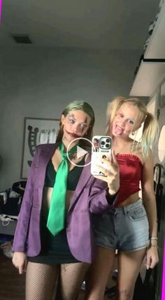 two women are taking a selfie with their cell phones in the room, one is wearing a purple jacket and the other has a green tie