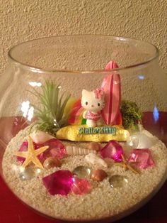 there is a glass bowl with sand and shells in it that has a hello kitty on top