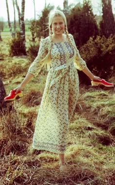 a woman in a long dress standing on the grass with red shoes and trees behind her