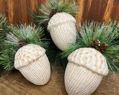 three white knitted acorns with pine cones and needles are sitting on a piece of wood