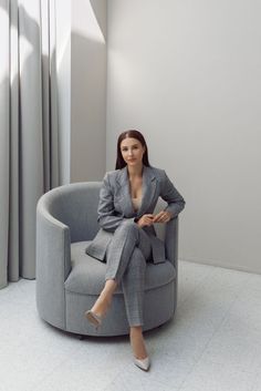 a woman sitting in a grey chair with her legs crossed, wearing a suit and heels