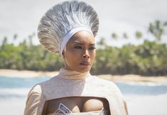 a woman wearing a white headdress standing in front of the ocean with palm trees behind her