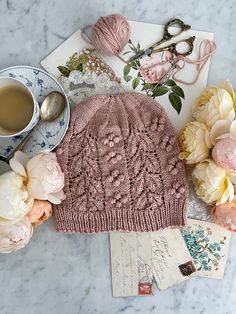 a pink knitted hat sitting on top of a table next to flowers and a cup of coffee