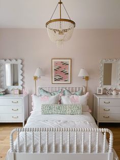 a white bed sitting under a chandelier next to a dresser with drawers and lamps