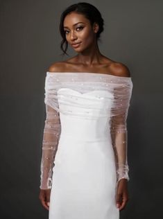 a woman wearing a white dress with sheer sleeves and pearls on the shoulders, standing in front of a gray background