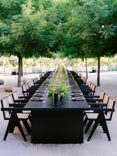 a long table set up with black chairs and greenery in the center, surrounded by trees