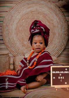 a baby sitting on the ground wearing a red and white striped dress with an orange beaded necklace