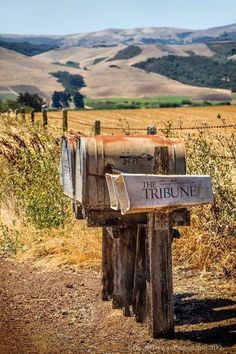 an old wooden mailbox sitting on the side of a dirt road next to a field