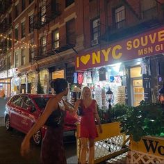 two women are standing in front of a store at night, talking to each other