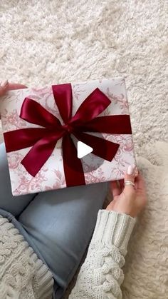 a woman holding a gift box with a red bow on it's ribbon and sitting on the floor