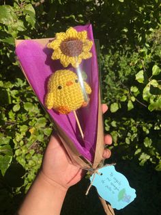 a hand holding a purple paper with a yellow crochet chicken on it and a blue tag