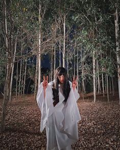 a woman dressed in white is standing in the woods with her hands up and two fingers out