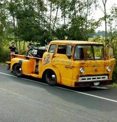 an old yellow tow truck is parked on the side of the road with two men standing next to it