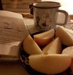 some apples are cut up on a plate next to an open book and a coffee mug