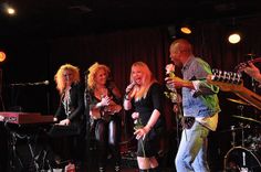 three women on stage singing into microphones with other people in the background at night