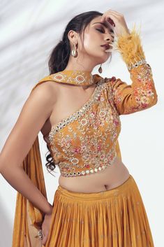 This gorgeous crepe and georgette mustard lehenga is pleated at the waist and tied together with a drawstring.  It is paired with a one shoulder printed blouse with beautiful mirror and handwork details and feather work on the sleeve hem.  The outfit is completed with a pre-pleated choker style dupatta with work details in the centre. Choker Dupatta Design, One Shoulder Ethnic Dress, Feather Blouse Designs, One Shoulder Dress Indian, One Shoulder Lehenga, Pleated Dupatta, Mustard Lehenga, Choker Dupatta, Pleated Lehenga